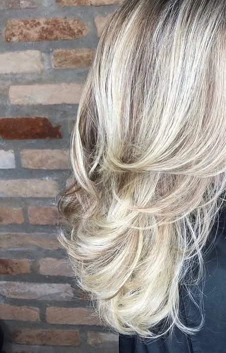 Cabelo loiro em camadas com movimento diante da parede de tijolinhos do salão; enquadramento sem rosto